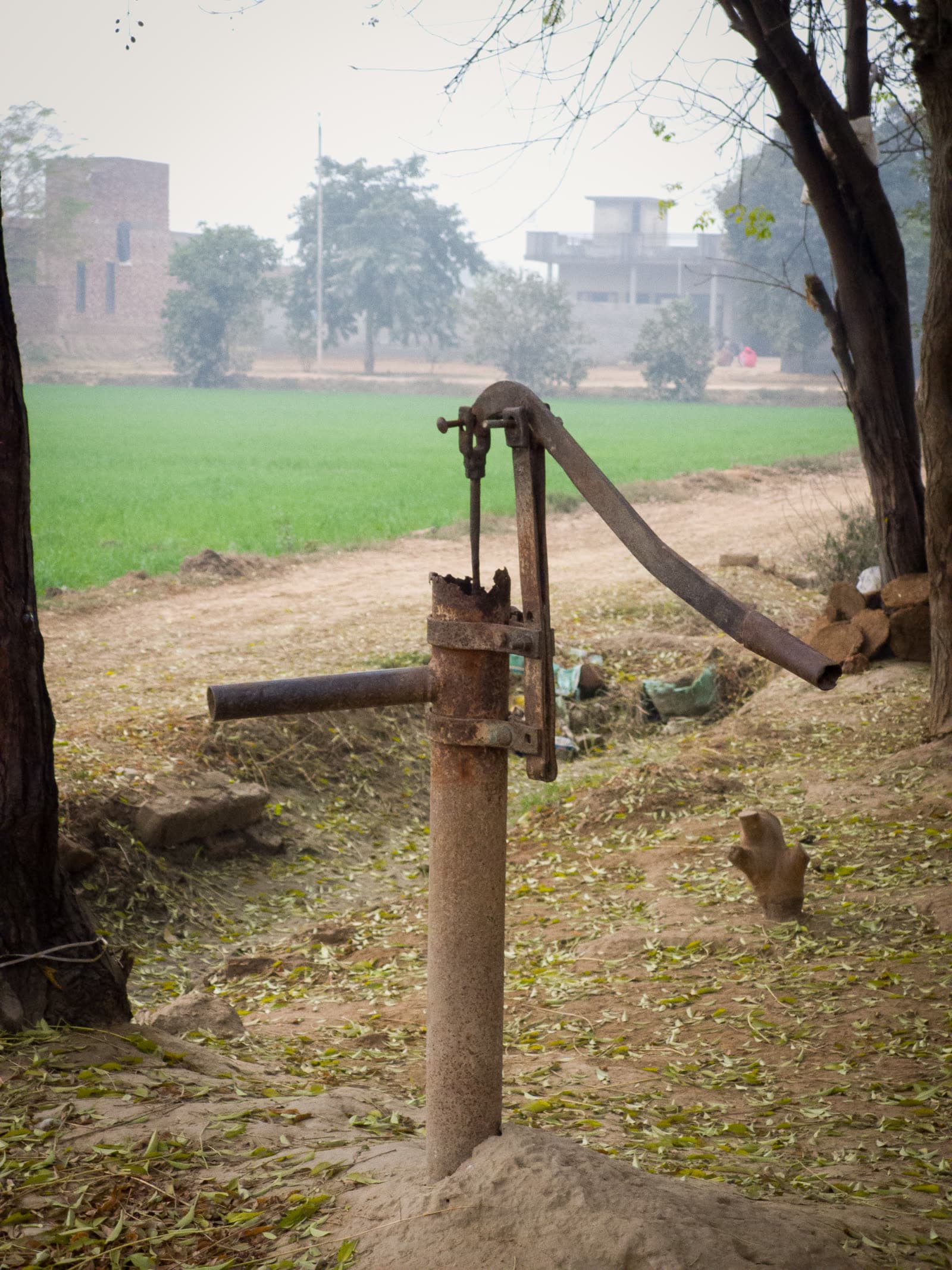 A rusted hand pump on a dirt path beside a green wheat field, with village houses in the haze behind